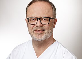 Mario Fassold Porträt eines Mannes mittleren Alters mit Brille und grauem Bart, in weißer medizinischer Kleidung, vor neutralem Hintergrund, mit freundlichem Ausdruck.