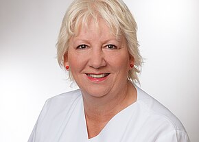 Sharon Porner Porträt einer lächelnden älteren Frau mit kurzem blondem Haar, roten Ohrringen und Lippenstift, in weißer medizinischer Arbeitskleidung vor neutralem Hintergrund.