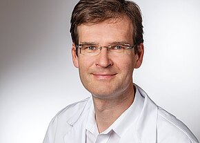 Micha Nowak Porträtfoto: Ein freundlich lächelnder Mann mit kurzer brauner Frisur und Brille trägt einen weißen Kittel und blickt in die Kamera vor neutralem Hintergrund.