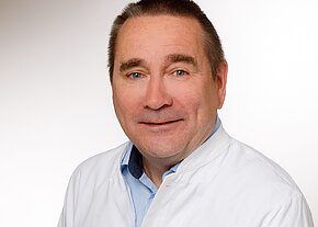 Dr. med. René Tandler Ein mittelalterlicher Mann mit einem freundlichen Lächeln trägt einen weißen Kittel über einem blauen Hemd. Er schaut in die Kamera und hat kurzes, leicht graues Haar. Der Hintergrund ist hell und unauffällig.