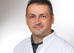 Aleksandar Cvetkovski Portrait eines Mannes in einem weißen Kittel. Er hat kurzes, dunkles Haar und einen freundlichen Gesichtsausdruck. Der Hintergrund ist hell und unscharf, wodurch der Fokus auf seinem Gesicht liegt.