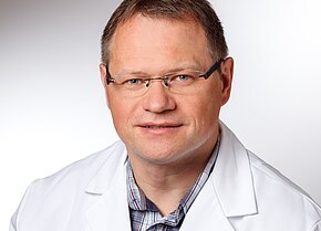 Dr. med. Rüdiger Heinz Porträt eines mittelalten Mannes mit Brille in weißem Kittel. Er blickt freundlich in die Kamera. Neutraler, heller Hintergrund.