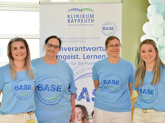 Vier Personen in hellblauen BASE-T-Shirts posieren vor einem Banner des Klinikums Bayreuth. Die Szene wirbt für die Ausbildungsstation BASE; freundliches Gruppenfoto bei einer Informations- oder Eröffnungsveranstaltung.