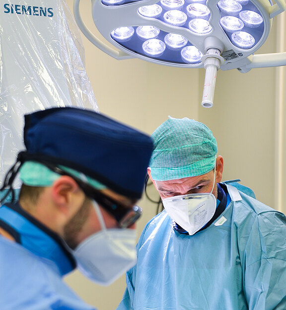 Zwei Chirurgen in OP-Kleidung arbeiten konzentriert im Operationssaal. Einer im Vordergrund unscharf, der andere blickt fokussiert auf das OP-Feld unter hellen Deckenleuchten.