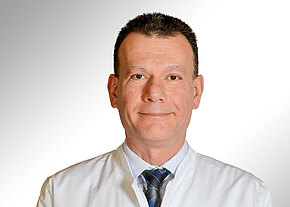 Dr. Alexander Bletkin Porträt eines Mannes in weißem Arztkittel und Krawatte, frontal aufgenommen vor neutralem Hintergrund, mit freundlichem, professionellem Gesichtsausdruck.