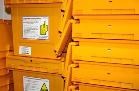 Mehrere stapelbare, orangefarbene Kunststoffbehälter stehen übereinander. Auf jedem Behälter sind Warnhinweise und Gebrauchsanleitungen in deutscher Sprache abgedruckt. Die Behälter sind für die Entsorgung von kontaminierten Materialien vorgesehen.