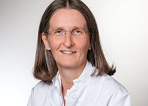 Heike Riedel Porträt einer erwachsenen Person mit schulterlangem braun-grauem Haar und Brille, freundlich lächelnd. Sie trägt einen weißen Arztkittel vor neutralem, hellem Hintergrund.