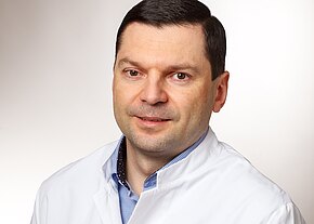 Vitaly Schur Porträt eines erwachsenen Mannes in weißem Kittel vor neutralem Hintergrund, leicht lächelnd, Blick zur Kamera.