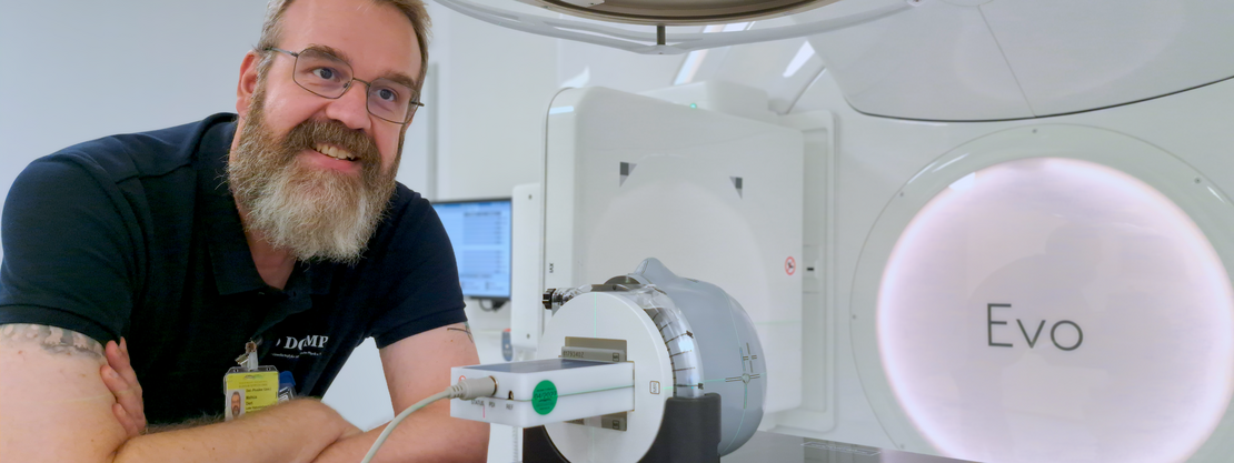 Ein lächelnder Techniker mit Bart lehnt an einem großen medizinischen Gerät. Auf dem Behandlungstisch ist ein zylindrisches Kalibrierungsgerät befestigt. Klinischer Raum mit Monitor im Hintergrund.