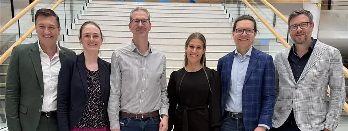 Sechs Erwachsene posieren lächelnd auf einer breiten Treppe in einem modernen Gebäude. Sie stehen nebeneinander, tragen überwiegend Business-Casual-Kleidung in unterschiedlichen Farben. Geländer links und rechts, helles Treppenhaus. Gruppenfoto, frontale Ansicht, entspannte Stimmung.