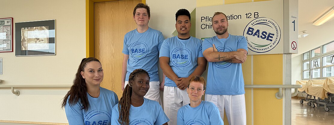 Sechs junge Erwachsene in hellblauen BASE-T-Shirts, teils in weißer Arbeitskleidung, posieren im Flur einer Krankenhausstation. Drei stehen, drei hocken davor. Im Hintergrund ein Stationsschild. Gruppenfoto eines Pflege- oder Ausbildungsteams auf einer Station.