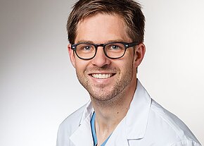 Dr. med. Dominik Schultes Porträt eines lächelnden Mannes mit Brille in weißem Arztkittel vor neutralem Hintergrund, Blick in die Kamera.