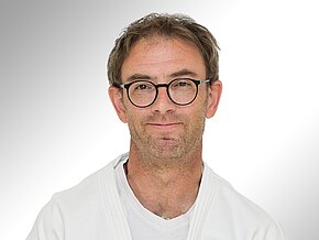 Dr. med. Christoph Erdmann Ein Mann mit Brille und kurzem, leicht zerzaustem Haar trägt einen weißen Judo-Anzug. Er lächelt freundlich in die Kamera. Der Hintergrund ist hell und neutral gehalten, was den Fokus auf sein Gesicht lenkt.