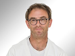Dr. med. Christoph Erdmann Ein Mann mit Brille und kurzem, leicht zerzaustem Haar trägt einen weißen Judo-Anzug. Er lächelt freundlich in die Kamera. Der Hintergrund ist hell und neutral gehalten, was den Fokus auf sein Gesicht lenkt.