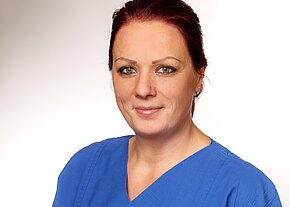 Sandra Bauer Porträt einer lächelnden Frau mit kurzen, roten Haaren, die ein blaues medizinisches Hemd trägt. Der Hintergrund ist hell und neutral, was die Aufmerksamkeit auf ihr Gesicht lenkt. Sie hat grüne Augen und wirkt freundlich und professionell.