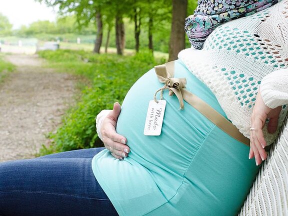 Schwangere Person sitzt draußen und hält den runden Bauch. Ein Schleifenband mit angeheftetem Schild liegt darüber; darauf steht „Made with love“. Kleidung in Türkis und Weiß.