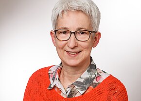 Uta Limmer Porträt einer lächelnden erwachsenen Person mit kurzem grauen Haar und dunkler Brille, vor neutralem Hintergrund. Sie trägt ein bunt gemustertes Hemd unter einem orangefarbenen Strickpullover, Blick zur Kamera.
