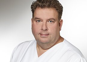 Stefan Scharnagel Porträt eines erwachsenen Mannes in weißer medizinischer Kleidung vor neutralem Hintergrund. Er blickt ruhig in die Kamera; Darstellung eines Gesundheitsmitarbeiters.