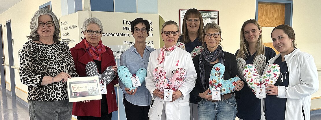 Gruppe im Krankenhausflur: Mitarbeitende und Unterstützerinnen, teils in Kittel oder Pflegekleidung, halten bunte herzförmige Kissen. Eine Person zeigt eine Urkunde. Die Szene zeigt eine Spende oder Übergabe von Trostkissen für Patientinnen auf einer Station.