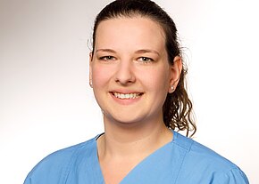 Julia Grasser Porträt einer lächelnden Frau in einem hellblauen medizinischen Kittel. Ihr Haar ist halblang und leicht gewellt, sie trägt kleine Perlenohrringe. Der Hintergrund ist hell und schlicht, was den Fokus auf ihr freundliches Gesicht lenkt.