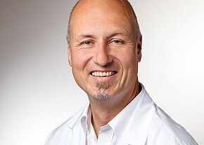 Dr. med. Christoph Dommke Freundliches Porträt: Ein glatzköpfiger Mann mit kurzem Kinnbart lächelt in die Kamera, trägt ein weißes Oberteil. Kopf-Schulter-Aufnahme vor neutralem Hintergrund, wirkt offen und professionell.