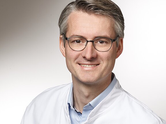 Porträt eines lächelnden Mannes mit Brille in weißem Kittel, blickt in die Kamera vor neutralem Hintergrund.