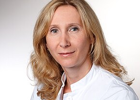 Verena Bauer Porträtaufnahme einer blondhaarigen Frau im weißen Kittel, neutraler Blick in die Kamera vor hellem Hintergrund.