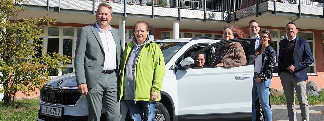 Sieben Erwachsene posieren vor einem mehrstöckigen Gebäude neben einem weißen Auto; eine Person sitzt am Steuer. Gruppenfoto bei Tageslicht.