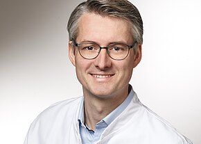 Univ.-Prof. Dr. med. Frank Kunath, FEBU, MHBA Ein Mann mit grauen Haaren trägt eine Brille und einen weißen Arztkittel. Er lächelt freundlich in die Kamera. Der Hintergrund ist hell und neutral.