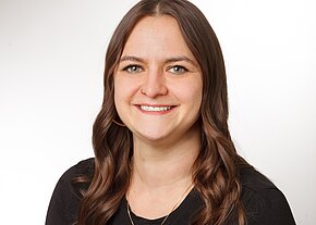 Nicole Spenner Porträt einer lächelnden Frau mit langen, gewellten braunen Haaren, die in die Kamera blickt. Neutraler Studiohintergrund, Oberkörper sichtbar, schwarzes Oberteil und dezenter Schmuck.