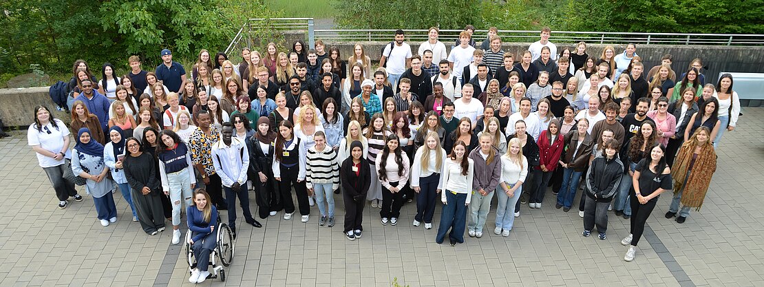 Großes Gruppenfoto vieler Menschen im Freien auf einem gepflasterten Platz, in mehreren Reihen angeordnet, zur Kamera gewandt; aufgenommen aus erhöhter Perspektive, im Hintergrund Bäume.