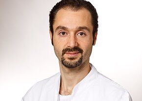 Alaa Fakhri Porträt eines Mannes mit dunklen, zurückgekämmten Haaren und kurzem Bart. Er blickt ruhig in die Kamera und trägt einen weißen Kittel. Heller, neutraler Hintergrund.