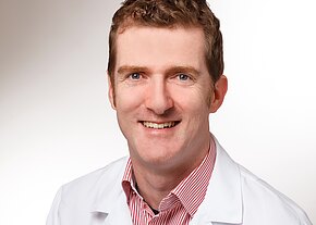 Univ.-Prof. Dr. med. Stefan Linsler Porträt einer lächelnden Person in weißem Kittel vor hellem Hintergrund, mit Blick in die Kamera. Unter dem Kittel ein rot-weiß gestreiftes Hemd.