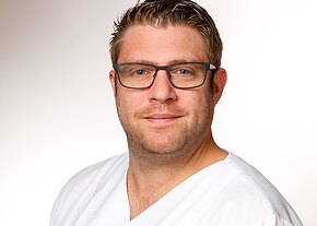 Station 24 Porträt eines Mannes mit Brille in weißer medizinischer Kleidung vor hellem Hintergrund; er blickt freundlich in die Kamera.