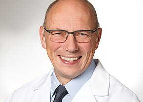 PD Dr. med. Gerhard Weyandt Porträt eines lächelnden, glatzköpfigen Mannes mit Brille in weißem Arztkittel, hellblauem Hemd und dunkler Krawatte vor neutralem Hintergrund.