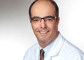 Prof. Dr. med. Alexander Kiani Porträt eines mittelalten Mannes mit Brille, der freundlich in die Kamera lächelt. Er trägt einen weißen Kittel, hellblaues Hemd und braune Krawatte. Neutraler, heller Hintergrund.