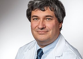 Dr. med. Claus Steppert Porträtfoto eines mittelalten Mannes mit grauem, lockigem Haar, der freundlich in die Kamera blickt. Er trägt einen weißen Arztkittel, ein hellblaues Hemd und eine dunkle Krawatte vor neutralem Hintergrund.