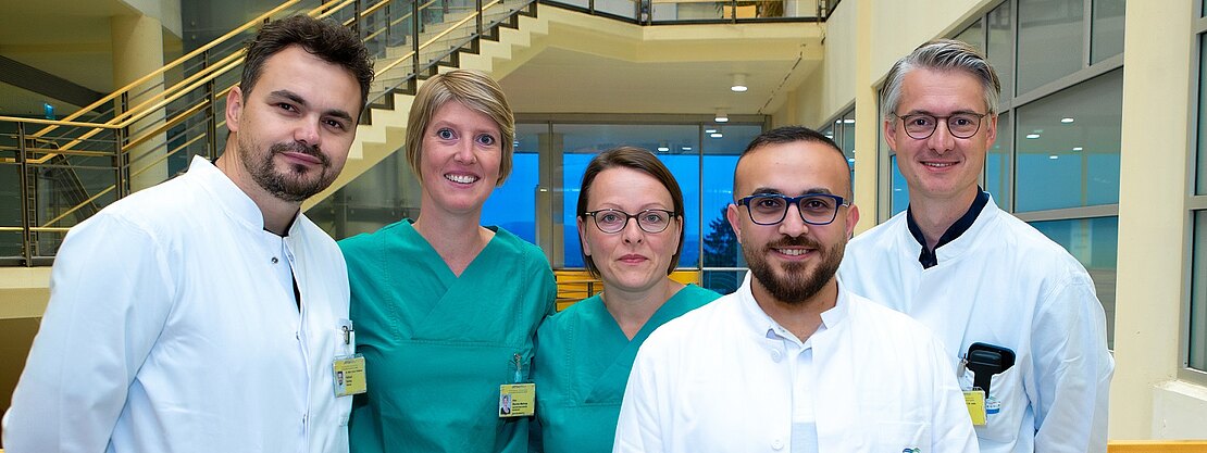 Fünf medizinische Fachkräfte stehen nebeneinander in einem hellen Krankenhausbereich. Drei tragen weiße Kittel, zwei grüne OP-Kleidung. Alle schauen in die Kamera, freundlich und professionell. Klinikumgebung im Hintergrund.