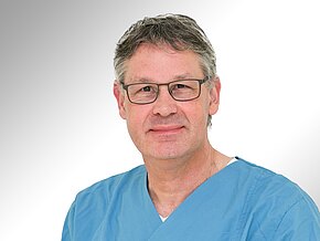 Jürgen Weigand Ein Mann mit grauen Haaren und Brille trägt einen blauen OP-Overall. Er lächelt freundlich in die Kamera. Der Hintergrund ist hell und neutral, was den Fokus auf seine Person legt.